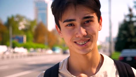 Shy-sincere-smile-of-asian-man-in-braces-looking-straight-at-camera,-showing-braces-while-standing-on-the-road-background,-urban-view
