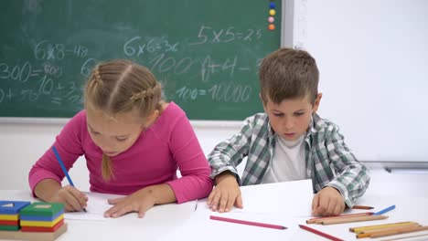 education-and-school,-happy-friends-classmates-sit-at-table-and-smilling-at-camera-on-background-of-blackboard