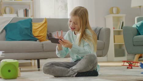 Smart-Cute-Girl-Sitting-on-a-Carpet-Playing-in-Video-Game-on-His-Smartphone,-Holds-and-Uses-Mobile-Phone-in-Horizontal-Landscape-Mode.-Child-Has-Fun-Playing-Videogame-in-Sunny-Living-Room.