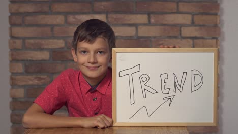 Kind-sitzt-am-Schreibtisch-mit-Flipchart-mit-Schrifttredstrend-auf-der-Rückseite-rote-Backsteinwand