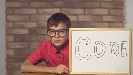 child-sitting-at-the-desk-holding-flipchart-with-lettering-code-on-the-background-red-brick-wall