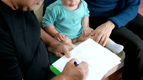 Attractive-baby-boy-draws-a-pen-with-his-grandparents-home-on-the-couch.-The-boy-stares-at-the-animals-that-drew-grandfather.-The-concept-of-different-generations