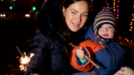 Young-mother-with-a-child-celebrate-the-new-year-on-the-street-with-a-sparkler.-Hugging-and-smiling-at-the-camera.-Against-the-background-is-decorated-Christmas-tree