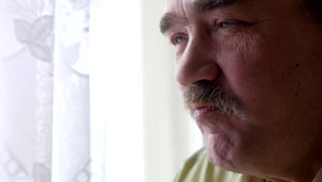 An-elderly-man-with-a-mustache-holds-a-cup-of-hot-drink-and-drinks.-He-sits-near-the-window-and-has-breakfast-at-home