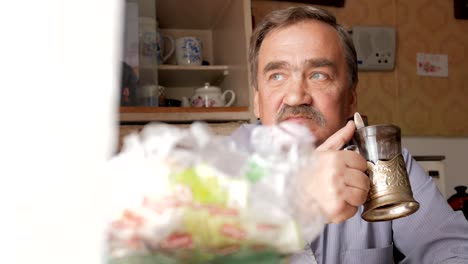 An-elderly-man-with-a-mustache-holds-a-cup-of-hot-drink-and-drinks.-He-sits-near-the-window-and-has-breakfast-at-home