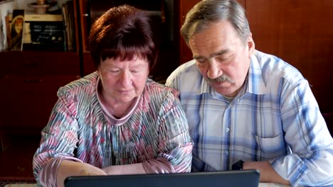 Una-pareja-de-ancianos-está-sentado-en-casa-en-la-computadora-portátil.-Una-mujer-lee-noticias,-un-hombre-con-un-bigote-se-sienta-junto-a-él-y-las-conversaciones