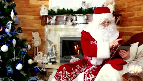 santa-using-tablet,-santa-claus-choosing-presents-with-the-help-of-gadget,-new-ideas,-fireplace