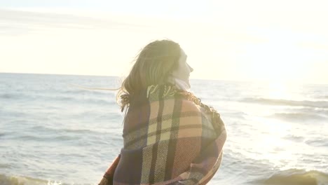 Young-beautiful-woman-raises-hands-up-with-plaid-on-the-shore-of-the-sea.-Happy-female-enjoying-the-sunset-on-the-beach