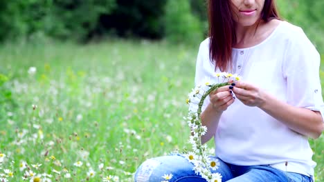 Verano,-en-medio-de-un-césped-de-la-manzanilla,-en-un-bosque,-una-mujer-joven,-una-morena-teje-una-guirnalda-de-chamomiles