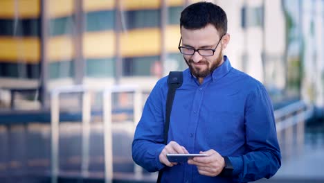 Fröhlicher-Mensch-Surfen-Smartphone-im-Büro.-Lächelnd-bärtigen-Büroangestellte-mit-Smartphone-am-Bürogebäude-auf-der-Straße