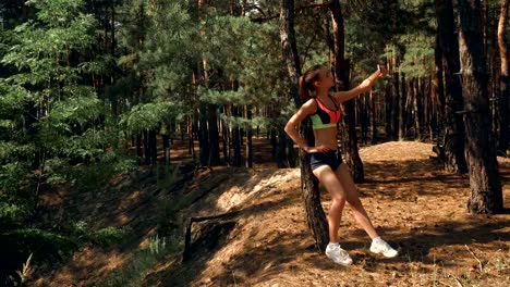 Athletic-beautiful-girl-in-short-shorts-and-top-stands-near-a-tree-and-makes-a-photo