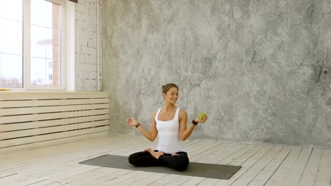 Beautiful-smiling-woman-holding-green-apple-sitting-on-exercise-mat