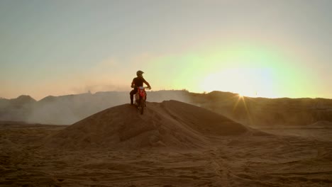 Largo-tiro-del-piloto-de-Motocross-extremo-en-fresco-casco-protector-de-pie-en-la-duna-de-arena-en-el-medio-escénico-mina-con-niebla-y-el-polvo-que-lo-cubre.