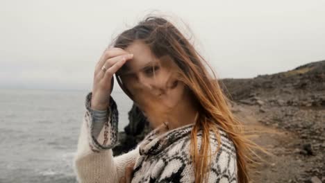 Portrait-of-young-happy-woman-standing-on-the-shore-of-the-sea-and-looking-on-camera,-smiling,-hair-waving-on-wind