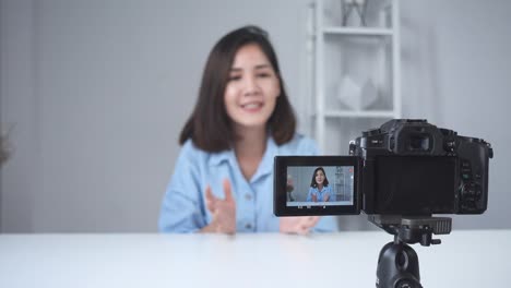 Glücklich-lächelnd-asiatische-Frau-oder-Beauty-Blogger-mit-Pinsel-und-Kamera-Videoaufnahme-und-winkenden-Hand-zu-Hause.-Videoblog-Blogging-Menschen-Schönheitskonzept.-Dolly-erschossen.