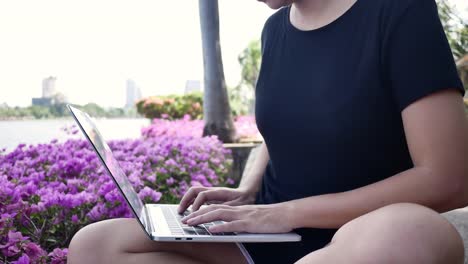 Schöne-asiatische-junge-Frau-sitzt-auf-der-Bank-im-Park-mit-Laptop.-Hübsche-Frau-auf-der-Suche-auf-ihrem-Computer-in-warmen-Frühlingstag.-Frauen-Lifestyle-Konzept.