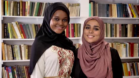 Porträt-von-zwei-charmante-junge-muslimische-Frauen-stehen-in-Bibliothek-und-lächelnd-in-der-Kamera,-afrikanische-Muslima-Hijab