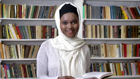 Young-african-muslim-girl-in-hijab-is-holding-book,-watching-at-camera,-religioun-concept,-booksheves-on-background