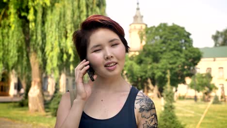 Portrait-of-young-asian-women-with-tattooed-hand-talking-on-phone-and-standing-near-university-in-park,-smiling