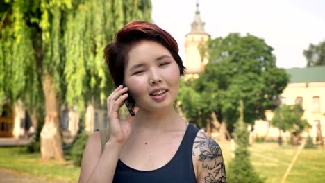 Retrato-de-jóvenes-mujeres-asiáticas-con-tatuada-mano-hablando-por-teléfono-y-de-pie-cerca-de-la-Universidad-en-el-parque,-sonriendo