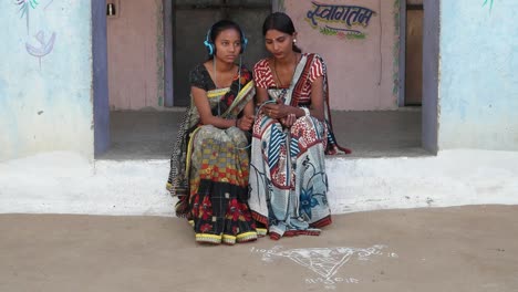 Dos-mujeres-disfrutando-escuchando-auriculares-música-en-teléfono-móvil-en-casa-rural-en-Rajasthan-India-con-ropa-tradicional-arte-impresión-brillante-colorido-diseño-artesanía-local-sari-porche-casa-interior-amor