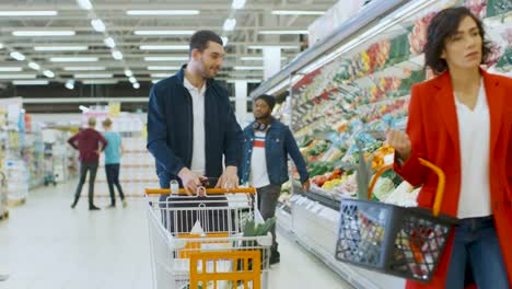 Im-Supermarkt:-gut-aussehender-Mann-mit-Smartphone,-schiebt-Einkaufswagen,-Spaziergänge-durch-frische-Produkte-Teil-des-Speichers,-wählt-einige-Produkte.-Andere-Kunden,-die-Produkte-zu-kaufen.