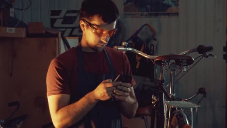 theme-small-business-bike-repair.-Young-caucasian-brunette-man-wearing-safety-goggles,-gloves-and-fartukhe-uses-mobile-phone-technology,-takes-notes,-checklist-in-bicycle-workshop