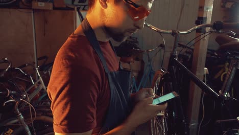 theme-small-business-bike-repair.-Young-caucasian-brunette-man-wearing-safety-goggles,-gloves-and-fartukhe-uses-mobile-phone-technology,-takes-notes,-checklist-in-bicycle-workshop