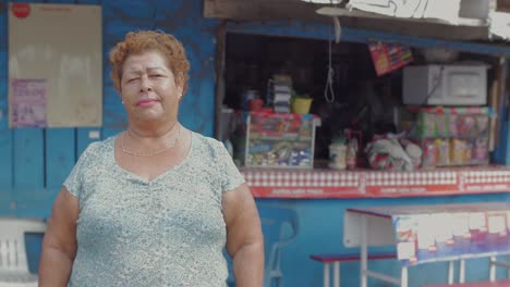 A-wide-portrait-of-an-older-hispanic-woman-looking-at-the-camera-standing-in-front-of-a-food-stand-in-Mexico