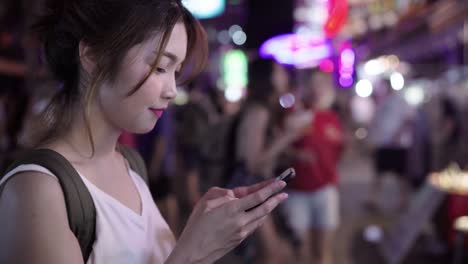 Asiatische-Frau-touristischen-Backpacker-lächelnd-und-mit-Smartphone-alleine-Urlaub-im-freien-unterwegs-Stadtstraße-in-der-Khao-San-Road-in-Bangkok-Bangkok---Thailand.