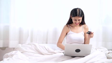 Frau-im-Bett-einkaufen.-Asiatische-Frau-mit-Laptop-sitzen-im-Bett-Shop-online,-gewinnt-einen-Preis-und-geben-Sie-Ihre-Kartennummer.-Online-shopping-Konzept.