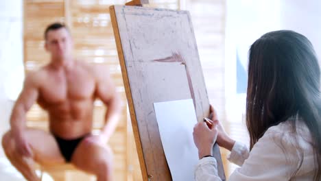 Young-woman-sits-behind-handsome-male-shirtless-model-and-drawing-him