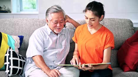 Daughter-reading-book-to-her-old-man-father-in-living-room.-Asian-senior-man-with-white-beard-and-asian-woman.-Senior-lifestyle-family-concept.