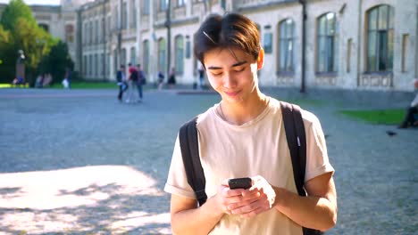 Hnsome-sonriente-a-asiatico-es-desplazamiento-su-teléfono-mientras-está-en-la-calle,-sunshines-anciant-edificios-detrás-de-él