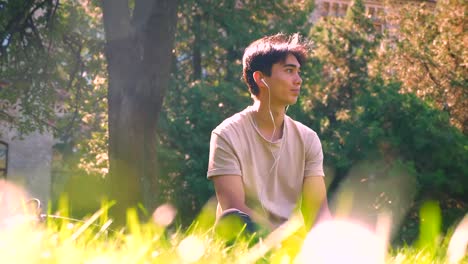 Quiet-asian-man-is-sitting-on-the-green-grass-under-the-tree-and-listening-to-music,-pure-nature-in-the-rush-of-big-city,-summertime