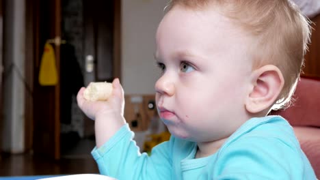 An-attractive-boy-2-years-old-eats-a-banana.-Sits-at-the-table-at-home.-Carefully-watching-cartoons-on-TV