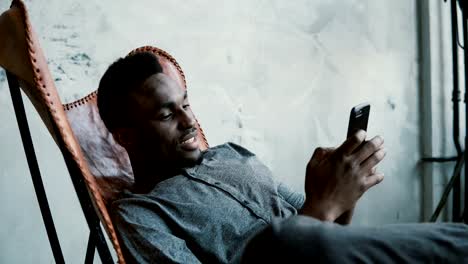 Portrait-of-African-man-sitting-in-chair,-using-a-Smartphone.-Handsome-male-smiles-and-looks-at-photos-in-his-phone