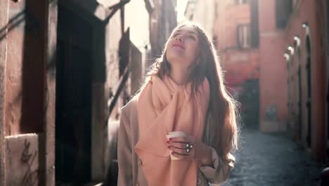 Portrait-of-young-smiling-woman-looking-at-camera.-Happy-brunette-girl-standing-in-morning-street-and-drinking-coffee