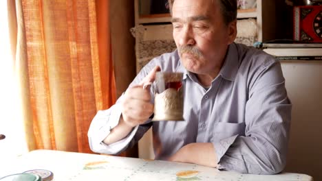 An-elderly-man-with-a-mustache-holds-a-cup-of-hot-drink-and-drinks.-He-sits-near-the-window-and-has-breakfast-at-home