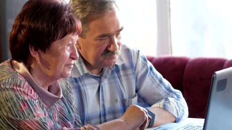 Una-pareja-de-ancianos-está-sentado-en-casa-en-la-computadora-portátil.-Una-mujer-lee-noticias,-un-hombre-con-un-bigote-se-sienta-junto-a-él-y-las-conversaciones