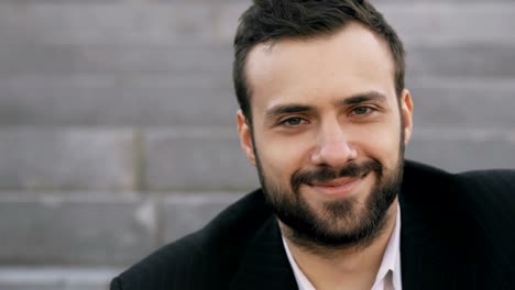 Close-up-portrait-of-bearded-young-businessman-smiling-and-looking-into-camera-in-street-outdoors