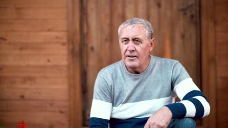 Old-man-with-gray-hair-sitting-in-front-of-camera-and-taking-conversation-with-somebody.-Wooden-background.-4K