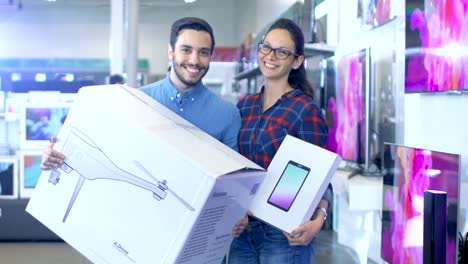 En-la-tienda-de-electrónica-feliz-pareja-joven/esposos-Poses-con-zángano-recién-comprado-y-Tablet-PC.-Tienda-es-grande-y-luminoso,-tiene-todos-los-últimos-televisores,-cámaras-y-teléfonos-inteligentes.