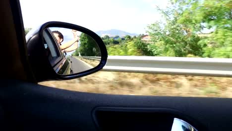 Rear-mirror-view-of-happy-young-girl-on-car-window-driving-on-the-road-having-fun-dancing-and-singing.-Party-concept