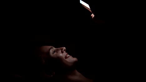 Young-smiling-woman-laying-on-a-couch-and-using-her-phone-in-a-dark-room,-chatting-in-the-darkness-with-smartphone,-communicating-with-friends-online