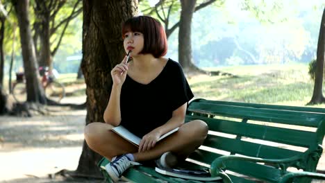 Happy-hipster-young-asian-woman-writing-into-her-diary-in-park.-Happy-hipster-young-asian-woman-working-on-notebook-in-park.-Student-studying-outdoors.-Lifestyle-woman-outdoor-concept.