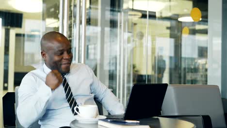 Cheery-African-American-businessman-smiling,-printing-and-working-on-his-laptop-in-glassy-cafe-during-lunch-break.-He-is-showing-yes-gesture-looking-happy