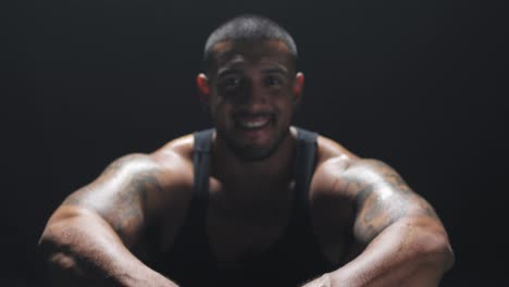 Zooming-shot-of-a-muscular-black-man-sitting-down-smiling-at-the-camera-on-a-dark-background