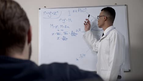 A-young-successful-African-scientist-in-a-white-coat-and-glasses,-stands-at-the-blackboard-with-formulas,-presentation,-lecture,-training-concept.-60-fps