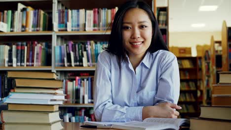 Porträt-des-jungen-schöne-asiatische-Studentin-mit-Haufen-von-Lehrbüchern-in-Bibliothek-Blick-in-die-Kamera-am-Tisch-sitzen.-Sie-lächelt-positiv.